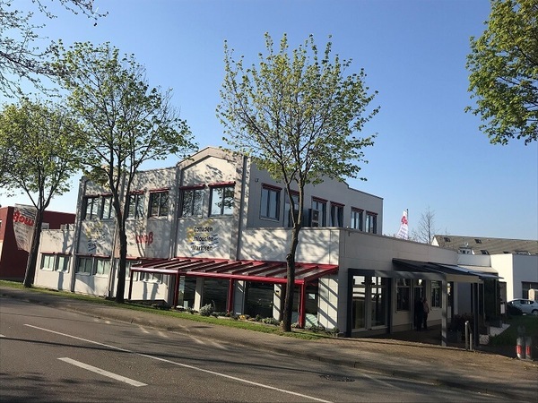 Modernes Firmengebäude mit großen Fenstern, Markisen und Bäumen an einer Straße.