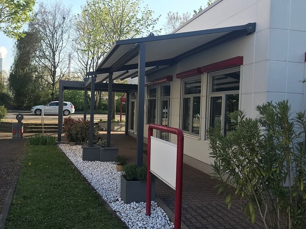 Moderne Terrassenüberdachung aus Aluminium vor einem Bürogebäude mit bepflanztem Vorgarten.