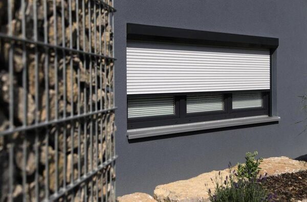 Modernes Fenster mit geschlossenem Rollladen an einer grauen Fassade und Steingabionen.