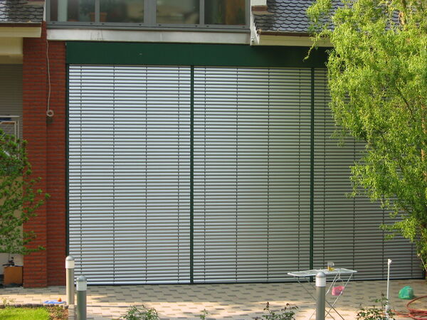 Große Aluminium-Rollläden vor modernem Haus mit Garten und gepflasterter Terrasse.